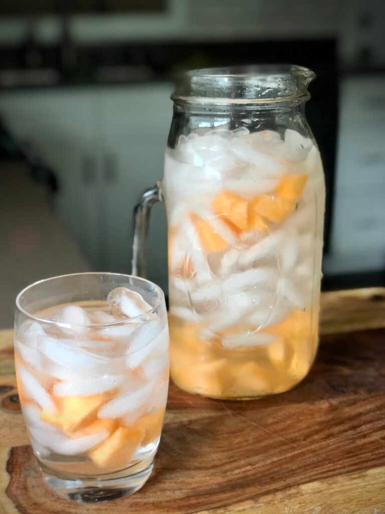 A large, glass pitcher and glass filled with ice and golden cantaloupe