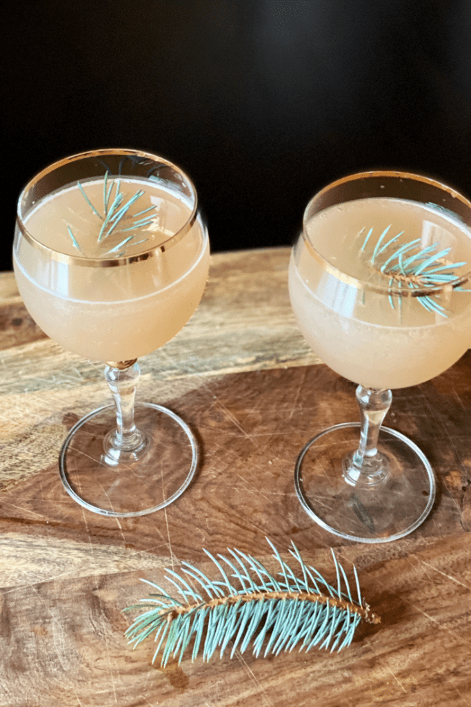 a lightly golden colored cocktail garnished with pine in a gold-rimmed glass